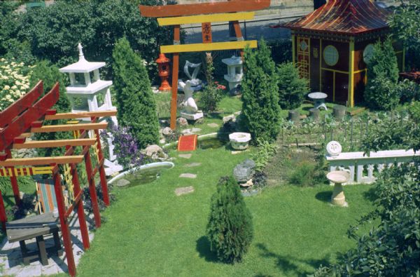 Elevated view of Sid Boyum's background taken from the second floor of his house. There is a large arch in the center with wind chimes hanging on it. Shrubs, trees, plants and other sculptures are on either side of a path, which is paved with stones spaced in the lawn. There are two ponds, one with a small arched bridge. There is a garden on the right. The Madison-Kipp Corporation factory is in the background.