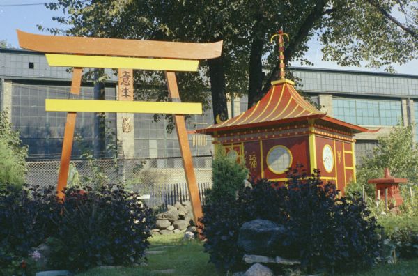 View of Sid Boyum's backyard, with an arch, pagoda, and sculptures among shrubs and trees. The Madison-Kipp Corporation factory is in the background.