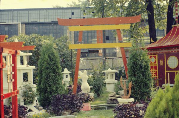 View of Sid Boyum's backyard. There is a large arch in the center with wind chimes hanging on it. Shrubs, trees, plants and other sculptures are on either side of a path, which is paved with stones spaced in the lawn. There are two ponds, one with a small arched bridge. The Madison-Kipp Corporation factory is in the background. 