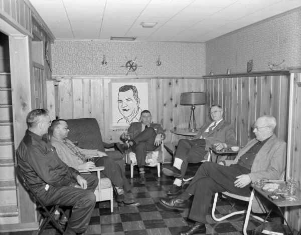 Portrait of a group of men attending a party in a basement. A poster with a drawing of a man is hanging on the back wall, with the title "Hugh Retirement." These are probably Gisholt employees.