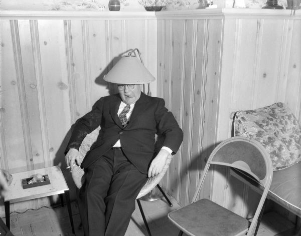 A partier at the "Hugh Retirement" party sitting in a chair with a lampshade on his head. He is sticking out his tongue and is holding a cigarette in his hand. These are probably Gisholt employees.