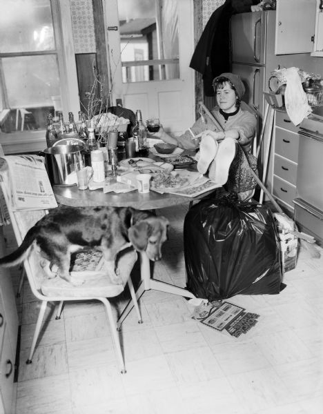 Inga sitting in a chair in a kitchen with her feet up on the table. She has a cigarette in one hand a drink in the other, and is wearing a scarf over curlers in her hair. A broom handle is resting on her lap, and there is a full garbage bag tied up on the floor. In the foreground a dog is standing on a chair. 