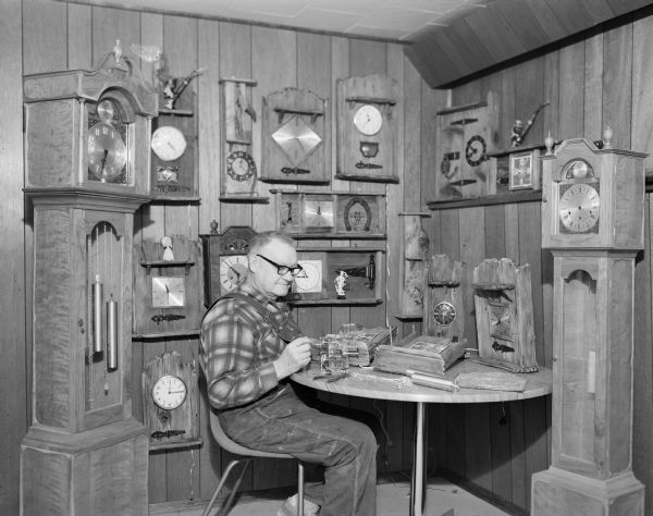 Walter Naef sitting at a table working on clocks. Clocks of many types and sizes, including grandfather clocks, are displayed around him.