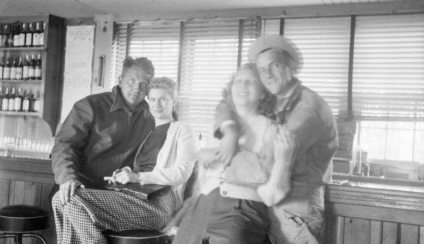 Group portrait, in a bar, of four people. On the right is a couple, a man standing with his arms draped around a woman in front of him. On the left is Sid with his sister, Bernice Boyum, who is sitting on a bar stool. 