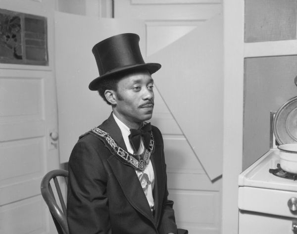 Waist-up profile portrait of John Cato, a Shriner, seated in Sid Boyum's kitchen. Cato is wearing a top hat, Shriner medallions, and a collar.