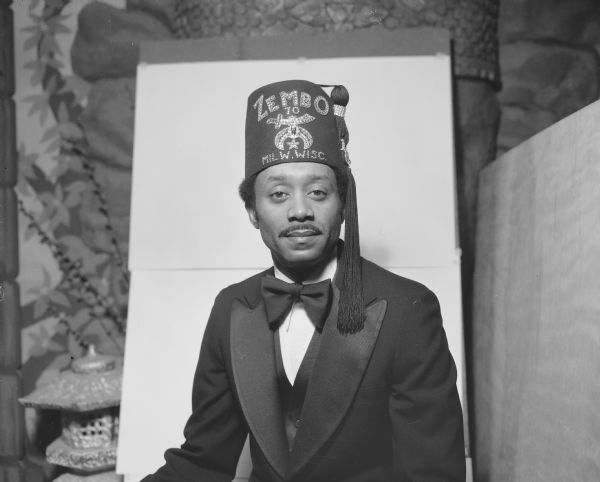 Portrait of John Cato, a Shriner, in Sid Boyum's front room, wearing a Zembo Temple fez and black formal suit. He is in front of a Buddha relief on the wall, and a small Japanese lantern.