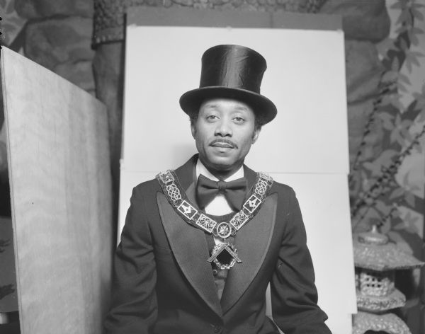 Portrait of John Cato, a Shriner, wearing a top hat, formal black suit and large chain necklace with a medallion. He is sitting in Sid Boyum's front room in front of Buddha head relief, with a small Japanese lantern.