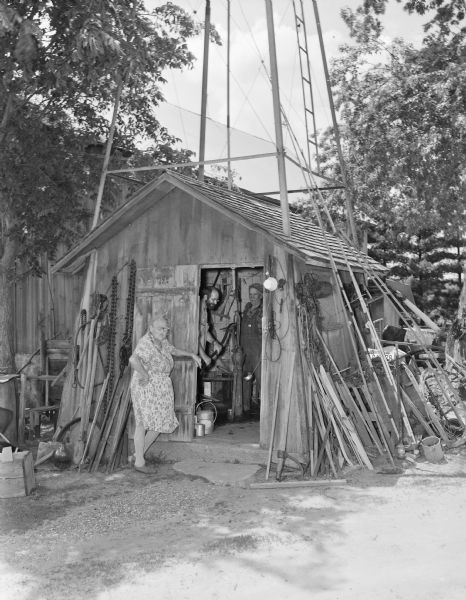 Reetz pump house, a small shed built under the base of a derrick. A woman is standing by the open door with her hand on her hip, and two men are inside, one of them probably Steve Hopkins, identified as an aide. One man is operating a hand pump. Gardening equipment and building material is propped against the shed.