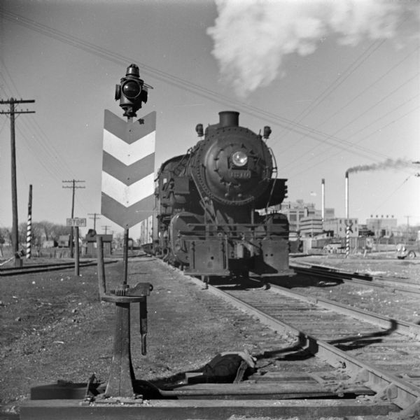 View alongside railroad tracks looking towards an approaching steam locomotive. In the background on the right is a power plant with smokestacks.