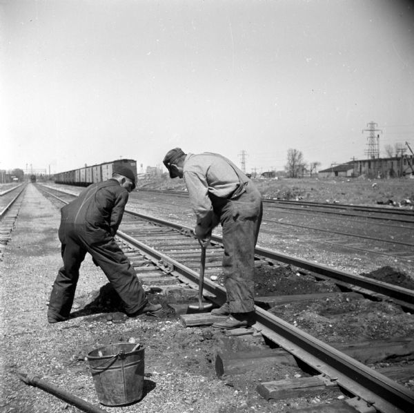 Two men, railroad workers, fixing the wooden railroad ties. In the background are railroad cars on another set of railroad tracks.m