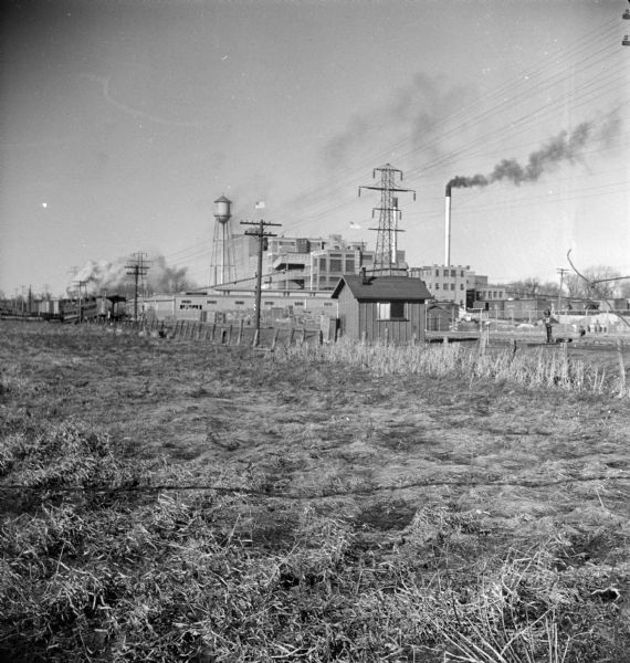 View across empty field towards railroad tracks. In the far background is a water tower next to a power plant.
