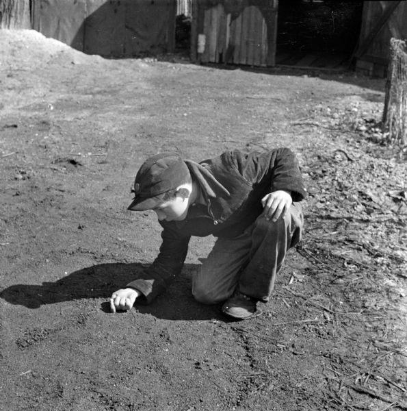 A boy is crouching on the ground playing a game of marbles outdoors in the dirt. In the background is a building with an open doorway.