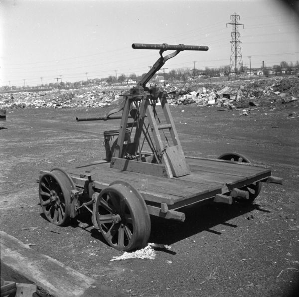 Rail handcar parked outdoors in a dirt yard. There is debris piled in the background, and in the far background are houses.