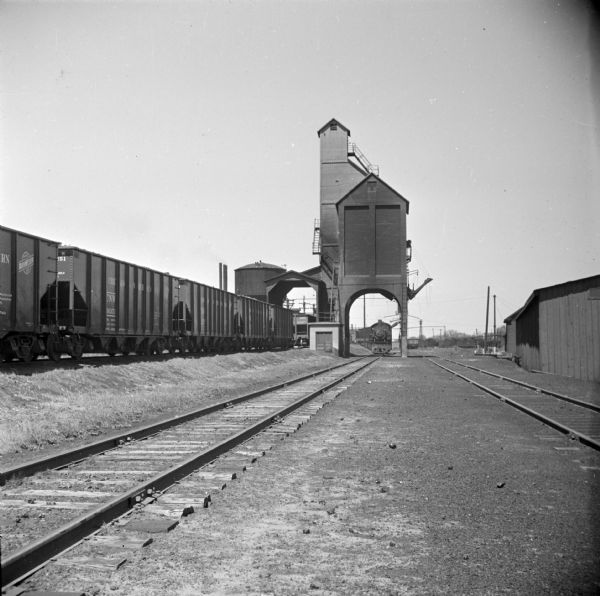 View alongside railroad tracks towards a railroad coaling tower. Railroad cars are on another set of tracks on the left. A locomotive is on the center set of railroad tracks.