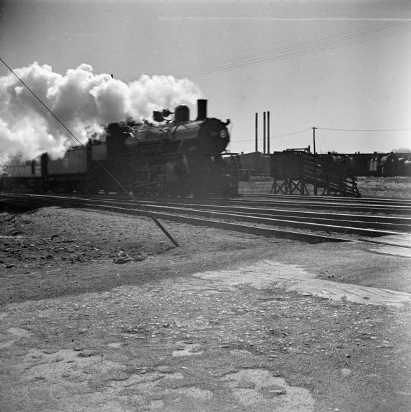 View towards a steam locomotive coming down one of a set of five railroad tracks at a railroad crossing. A group of locomotives are parked in the background near industrial buildings.