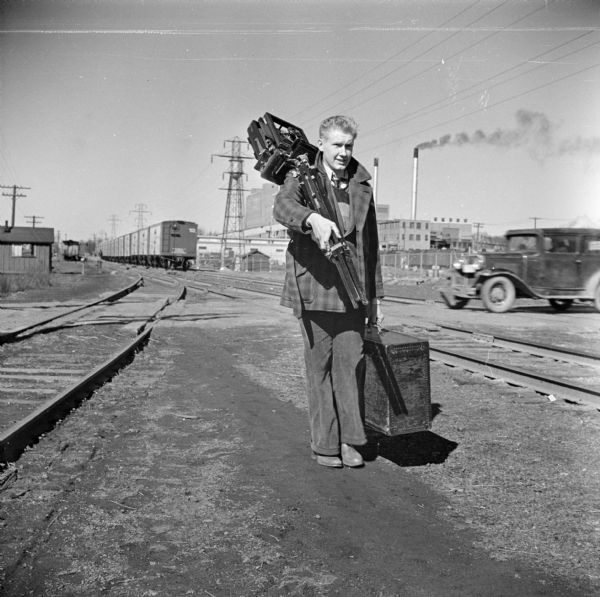 A photographer is walking near railroad tracks at a railroad crossing, with an automobile at the crossing in the background on the right. The photographer is carrying professional photography equipment, including an 8x10 field camera. In the background is a power plant.