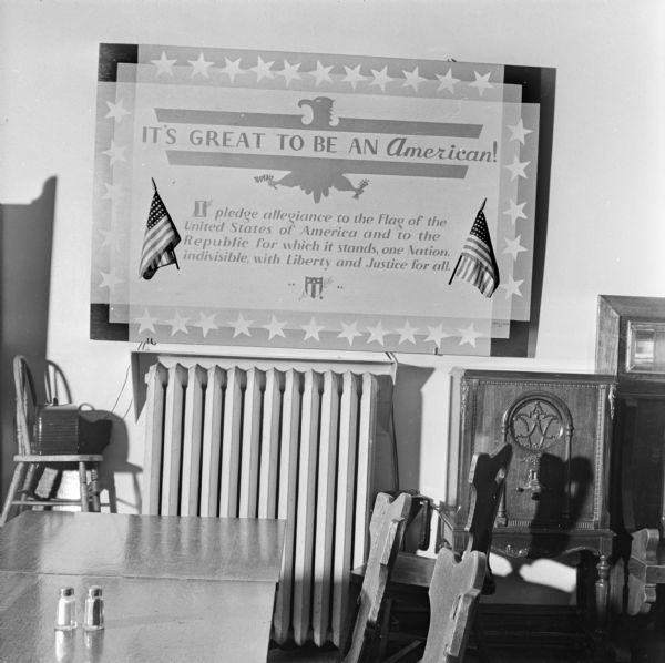 A sign titled: "It's great to be an American!" includes an eagle symbol, American national flags, and the Pledge of Allegiance, hanging on the wall inside a room. 