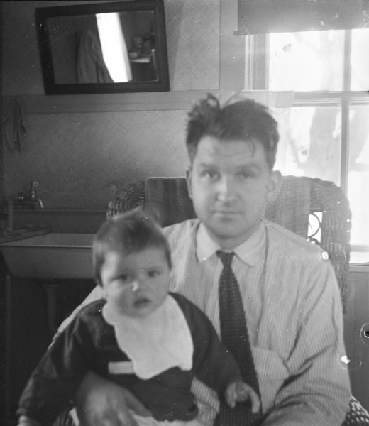 Indoor portrait of Sid sitting with his son, Steve, in his lap. Behind them is a sink near a window. 