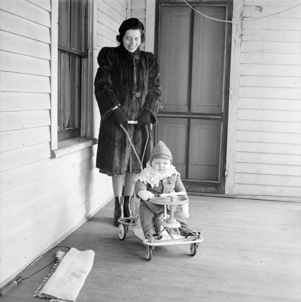 Portrait of Margaret Boyum, wearing a fur coat, standing on a porch holding the handle of a Taylor Tot stroller that her son, Steve Boyum, is sitting in. 