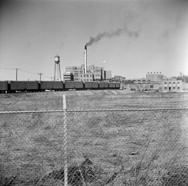 View over chain link fence towards a train on the railroad tracks near a power plant and water tower. 