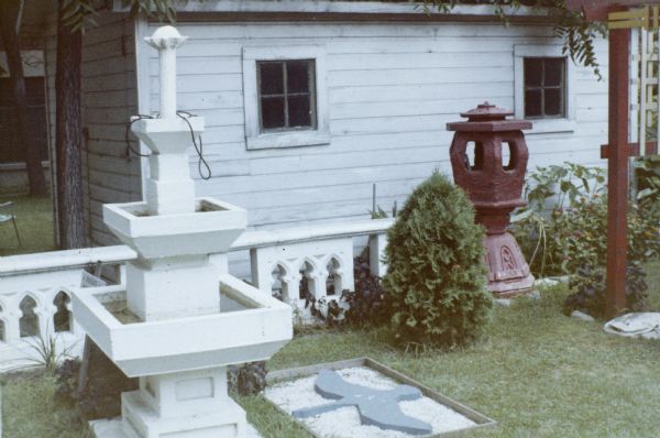 "Six-Sided Red Lantern," "Three-Tiered Fountain" and a flat goose cut from gray stone (laid in a bed of white gravel) in Sid's backyard. A corner of the painted torii gate is on the far right. On the other side of the fence in the background stands a garage. 