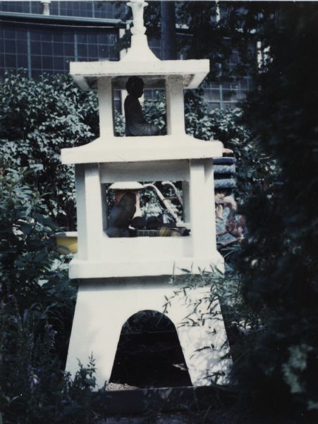 Sid's lantern sculpture with a Buddha sculpture inside is surrounded by trees and plants. The sculpture is called "Three-Tiered Tower." The Madison-Kipp Corporation factory building is in the background. 