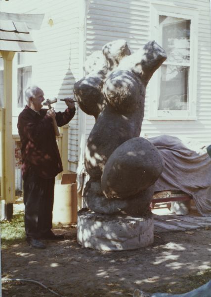 Sid standing and carving the sculpture of a woman's torso "Biomorphic Female Abstract in Pink" with a hammer and a chisel in his backyard.