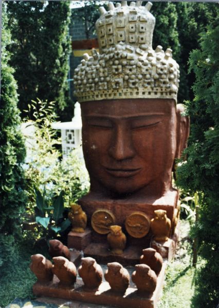 Buddha head, titled "Monumental Oriental Head," surrounded by trees and shrubs in Sid's background. With dimensions of 110 x 45 x 78 inches, it sits on a thick base ornamented with medallions bearing symbols of the Zodiac. Figural owls sit beneath the bust on the base and in the space in front. The white "Lantern in Beige" is in the background.