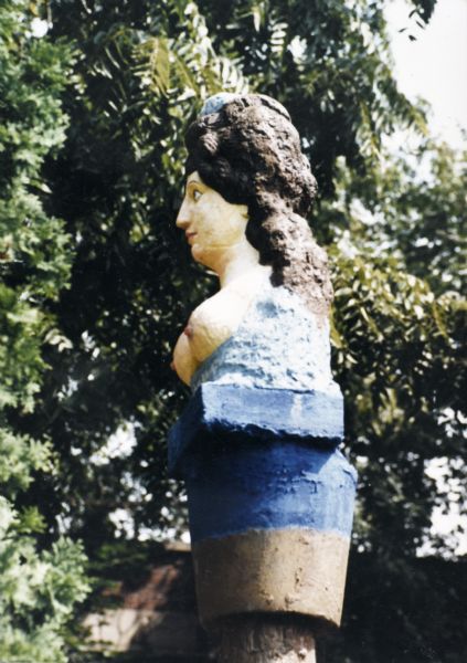Profile of the concrete-cast sculpture,"Feminine Portrait," in Sid's backyard. The woman is bare breasted and has thick brown hair with a blue ornament on the crown of her head. The bust sites on a square blue base supported by a column of the same color. This sculpture resembles a figurehead on the prow of a ship.