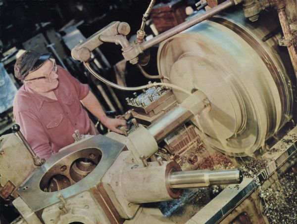 Color illustration art of a machine operator working on a six-arm turret. He is wearing a billed cap and safety goggles. 