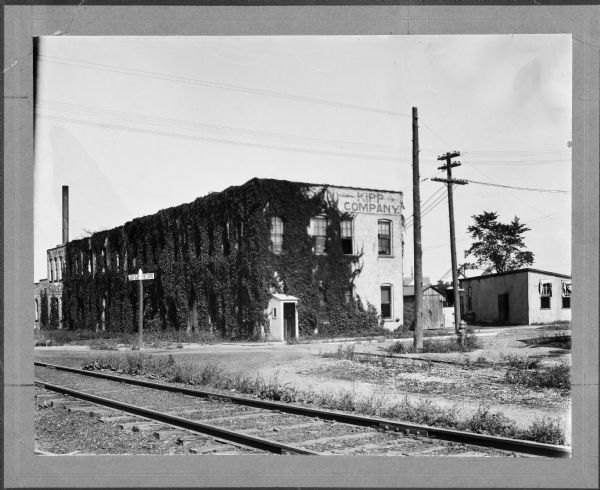View towards the building of Plant No. 1 of the Madison-Kipp Corporation on the other side of Waubesa Street. Ivy covers its brick walls. Railroad tracks run diagonally in the foreground and a smokestack is in the background. Text on the street sign at the tracks reads: "Look out for cars."
