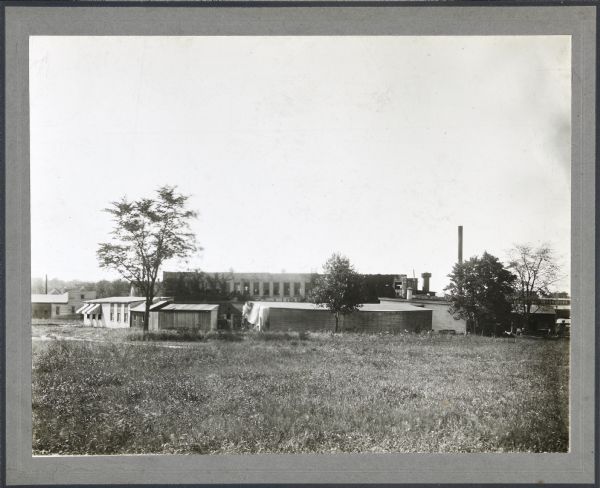 View across field towards structures in front of the Madison-Kipp Corporation building, pictured in the background. Its smokestack is visible to the right of the two-storied building. Trees dot the undeveloped landscape. 