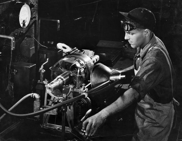 Illustration art of a machine operator in work clothes and a billed cap with safety goggles standing next to a Type S Dynetric Balancer at the Howell Electric Motors Company. The balancer was one of the new products manufactured by Gisholt Machine Company.