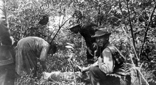 An American Indian woman is bending over the carcass of a young deer on the ground in a wooded area. A man, far right, is looking at the camera while holding the deer's ear. He has a pair of binoculars slung over his shoulder.  Two other men are watching.