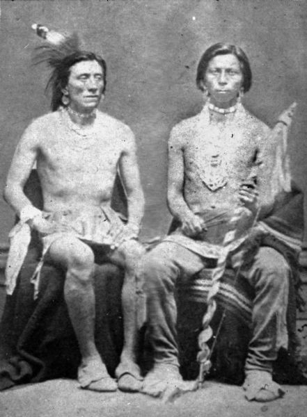 Two unidentified Native American men, probably Ho-Chunk, posing for a seated, full-length studio portrait. Each is seated on a blanket and is shirtless. The man on the left wears a breechcloth, flapped moccasins, necklaces and earrings. He has a small headdress. The man on the right wears a breechcloth with leggings, moccasins, elaborate necklace and earrings. He holds a staff in his left hand.