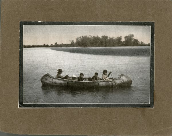 View across water towards an Ojibwe couple paddling a birch bark canoe with their five children seated in the center. The family is dressed in western style clothing. The father, sitting in the rear, and one of the children are wearing hats.