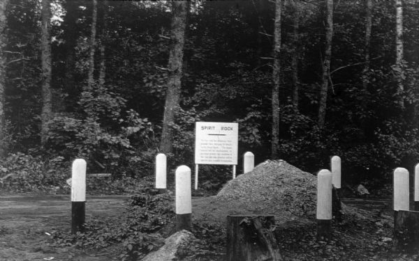 View of a sign identifying the Spirit Rock, which is a large boulder surrounded by sturdy protective posts. There are tree stumps in the foreground and a wooded area behind the sign.  