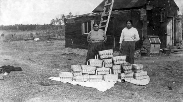 Two unidentified Ho-Chunk women posing outdoors behind a pile of baskets made from split black ash. There is a tarpaper covered dwelling behind them with a doghouse in front. A ladder leans against the side of the building. The woman on the left wears a print blouse and print skirt, a common style for Ho-Chunk women. The woman on the right wears a plain skirt and blouse.