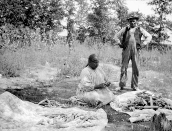 A Native American woman is sitting on the ground preparing corn for drying. A man, with arms akimbo, is standing nearby, observing. Husked corn is piled on a patchwork quilt in the foreground.
