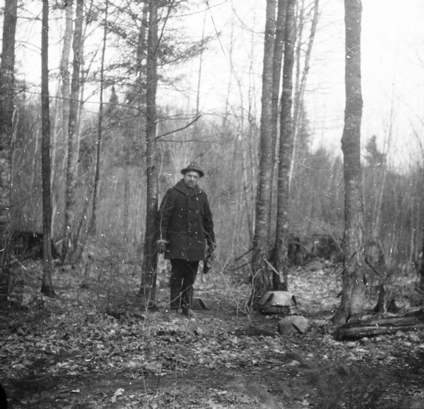 An unidentified man wearing a hat and double breasted overcoat is holding a binocular case in his left hand and posing beside maple trees that have multiple spiles, or taps, in their trunks. There are maple syrup buckets on the ground.