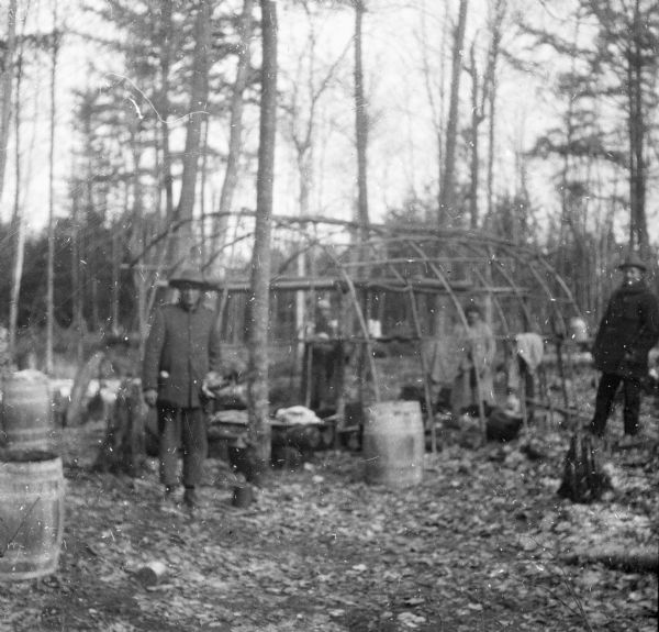A Native American man, left, posing in front of the sapling frame of a shelter at a maple sugar camp. Two Native American women are standing under the framework, and a man of European descent is at standing on the far right. Several wooden barrels and small buckets are on the ground.