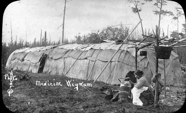 A Ho-Chunk woman with two children, a toddler who is standing, and an infant on a cradleboard, beside a long lodge (ciiserec). The lodge is covered with cattail reed mats. The woman is wearing a printed blouse, and an apron over a long skirt. Two kettles are hanging from a pole framework in the foreground. "Medicine Wigwam" and "Hoover Photo" are printed on the photograph.