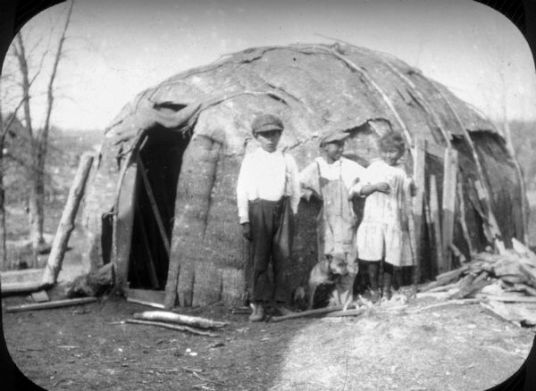 Three Native American children, two boys and a girl, posing with their dog in front of a wigwam. The children, two boys and a girl, are wearing European-style clothing. The boy in the center is wearing bib overalls, and both boys are wearing caps.