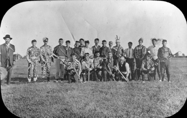 Outdoor group portrait of a Native American lacrosse team posing holding their sticks before a match. A man holding a drum is standing on the far left. Two players with round ornaments attached to their shirts and pants are standing on the left side of the group. Most of the other players are wearing subdued, European-style clothing, but some are wearing boldly patterned shirts, one with stars and stripes.  Several of the men are wearing neckties or hats, and one is wearing a small feathered headdress.