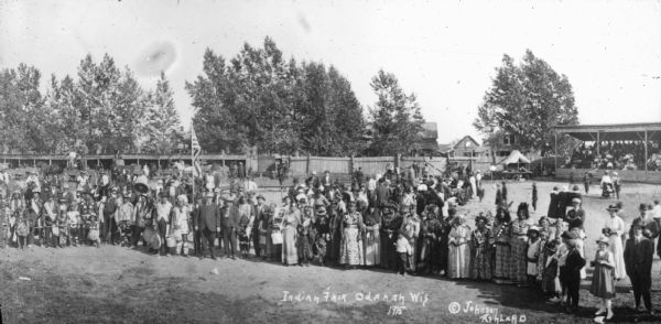 Elevated group portrait of men, women and children posing at the fairgrounds at Odanah. Most of the people are Native Americans, probably Ojibwe, with a number of white attendees. Left of center, a man is holding an American flag on a pole. There are horse stalls and horse-drawn wagons in the background, and a grandstand on the right. A house and barn are beyond the stockade-style fence.