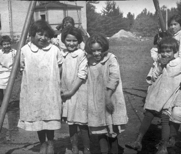 A group of eight Native American school girls posing near playground equipment. A brick school building is in the background.  The girls are dressed in identical uniforms, with smocks or jumpers over their dresses. 