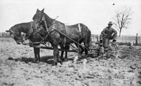 An unidentified Native American man posing while sitting on a corn planter pulled by a team of horses.
