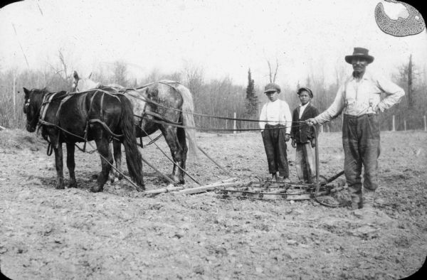 An Ojibwe man is standing and holding the reins of his team of two horses hitched to a harrow. Two boys are standing behind him. In the background are leafless trees beyond the field.
