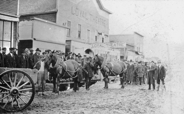 View across unpaved road towards a Native American man wearing a plaid overcoat with two work horses. A crowd of men are watching from the elevated wooden sidewalk and from the street, and in the background is the Loyal Theater. A Caucasian man is on the far right, possibly Joseph Wojta, pointing toward one of the horses. Wojta was a University of Wisconsin Extension agent who promoted improved farming techniques among Wisconsin's various Indian tribes.