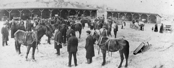 Elevated view of a Native American man holding the lead of a horse as it is evaluated by another man, possibly Joseph Wojta, who is wearing a hat and long overcoat. There is a crowd of onlookers, including Native American women wearing blanket shawls, as well as men and children. In the background are two open buildings where wagons are parked in stalls.  